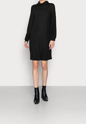 Schwarzes Langarmkleid mit Wasserfallausschnitt, aus einem weichen Stoff; knielang mit einer lockeren Passform; kombiniert mit schwarzen, hochhackigen Stiefeletten.
