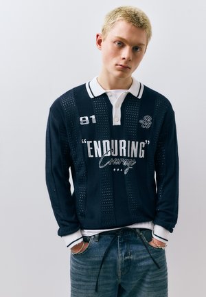 Junge Person mit kurzen blonden Haaren, trägt ein marineblaues Hemd mit weißem Kragen, darauf der Schriftzug "ENDURING Courage", und blaue Jeans, Hände in den Taschen, vor einfarbigem Hintergrund.