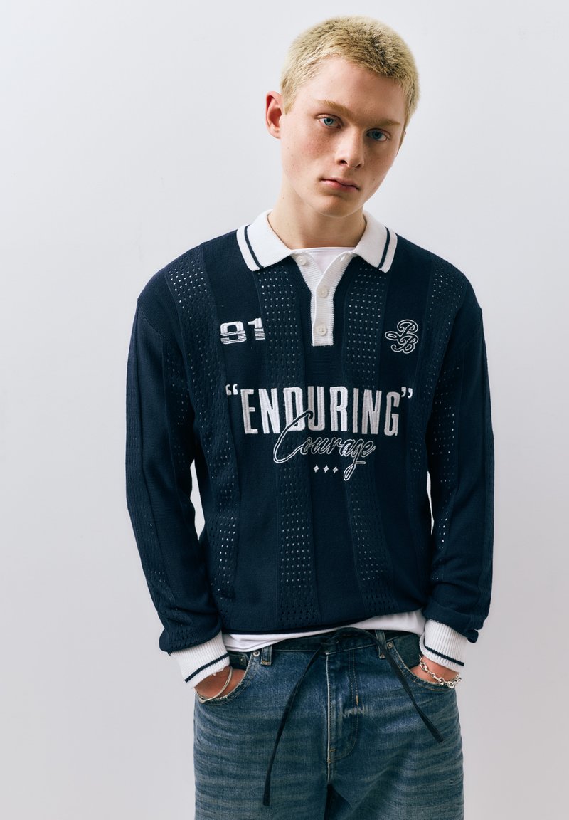 Jeune personne aux cheveux blonds courts portant une chemise bleu marine avec un col blanc, un texte "ENDURING Courage", et un jean bleu, les mains dans les poches, sur un fond uni.