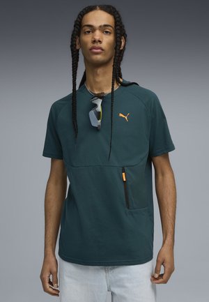 Nuori mies pitkillä letillä, päällään tumma vihreä Puma-paita, jossa on oranssi logo, ja aurinkolasit kaulassa, vaaleansiniset farkut.
