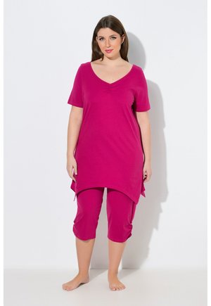 Donna in piedi a piedi nudi, indossa una tunica rosa acceso a maniche corte e pantaloni corti abbinati, su sfondo bianco.