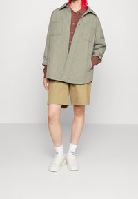 Olivgrön, vadderad jacka, brun ribbad skjorta, khaki shorts, vita strumpor och vita sneakers, står mot en ljusgrå bakgrund.