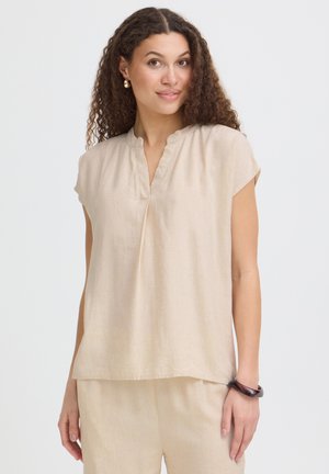 Vrouw met krullend haar die een beige blouse met korte mouwen en bijpassende broek draagt, geaccentueerd met gouden oorbellen en een donkere armband.