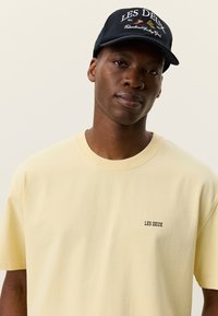 Απαλό κίτρινο βαμβακερό μπλουζάκι με στρογγυλή λαιμόκοψη, κοντά μανίκια και μικρό μαύρο λογότυπο "LES DEUX" στο στήθος. Μαύρο καπέλο με λεπτομέρειες λογότυπου.