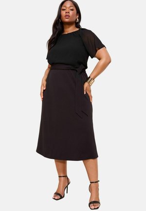 Blouse en chiffon noir à manches courtes associée à une jupe portefeuille noire taille haute. Le modèle porte des talons strappy noirs et des accessoires en or.