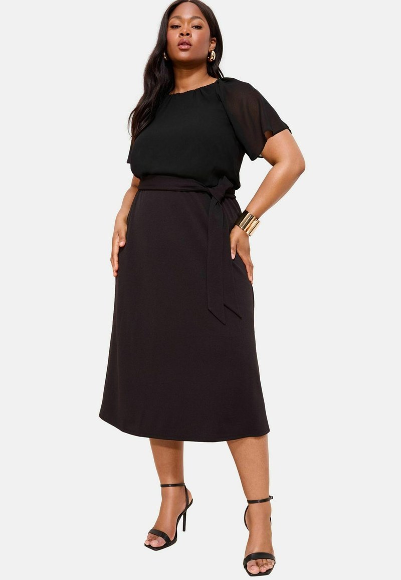 Schwarze Chiffon-Bluse mit kurzen Ärmeln, kombiniert mit einem schwarzen, hochgeschnittenen Wickelrock. Das Modell trägt schwarze Sandalen mit Riemchen und goldene Accessoires.