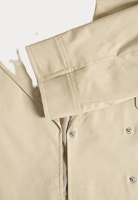 Beige jas met een kraag, voorzien van een knoopsluiting en zilveren accenten. Het materiaal is glad met gestructureerde naden en een strak ontwerp.