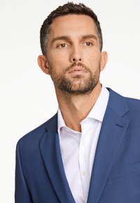 Granatowa marynarka o gładkiej fakturze, z klapami typu notch i jedną kieszenią na piersi, noszona na białej koszuli z guzikami.