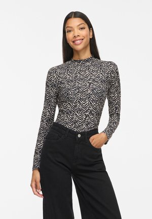 Femme aux longs cheveux foncés souriant, portant un haut à manches longues à motif zigzag noir et blanc et un pantalon noir taille haute, une main dans la poche.