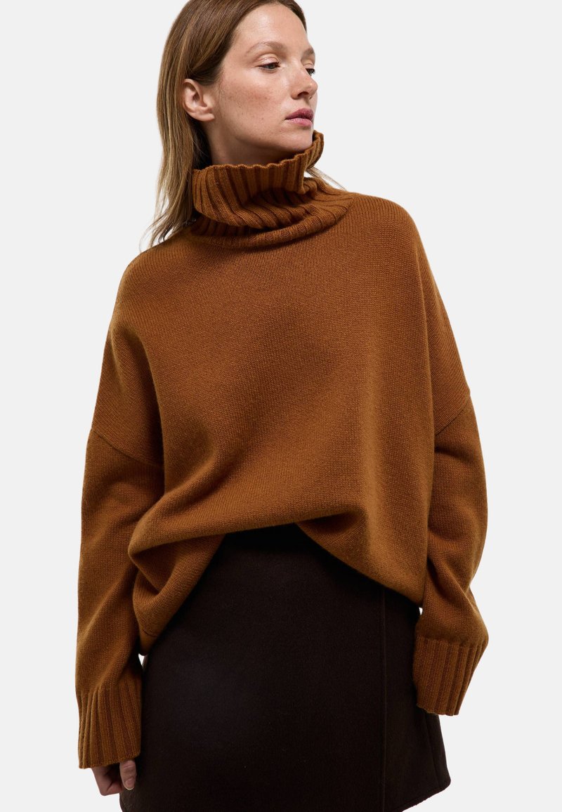 Pull en maille oversize marron avec col roulé, présentant des poignets et un col côtelés, associé à une jupe structurée noire.