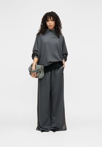 Γκρι φαρδιά φούτερ με ψηλό λαιμό και μακριά μανίκια, συνδυασμένο με παντελόνι φαρδιάς γραμμής στο ίδιο σχέδιο. Το μοντέλο κρατάει μια τσάντα από υφή υφάσματος.