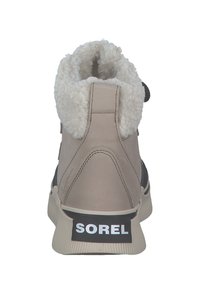 Beige schoen met een zachte witte kraag, gestructureerd oppervlak, zwarte accenten en een opvallend "SOREL"-logo op de hiel. Waterdicht ontwerp.