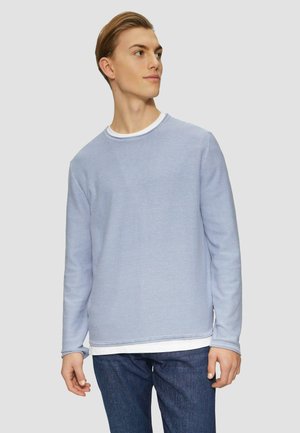 Lichtblauwe long sleeve trui van zachte stof met geribde halslijn en mouwen, gelaagd over een wit shirt, gecombineerd met donkere spijkerbroek.