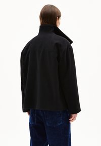 Schwarze Wollmischjacke mit hohem Kragen und lässiger Passform, mit einer geraden Rückennaht. Getragen über blauen Cordhosen.