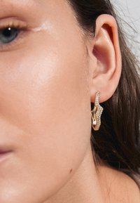 Orecchini d'oro con un design ondulato, caratterizzati da strass trasparenti su un lato. Vista ravvicinata che evidenzia la texture e i dettagli intricati.