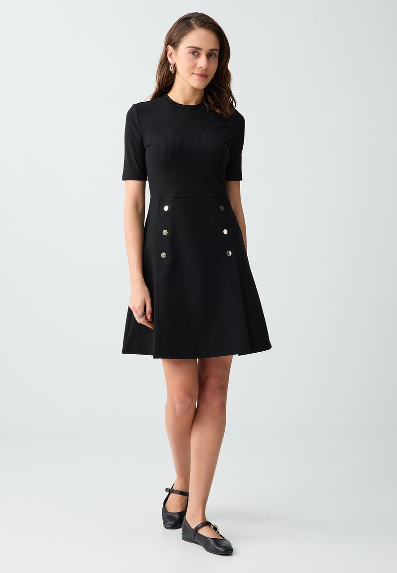 Jeune femme portant une robe noire à manches courtes avec des boutons métalliques sur la jupe et des chaussures plates Mary Jane noires, debout devant un fond uni.