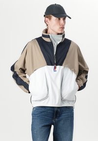 Fargeblokkert vindjakke i beige, marineblå og hvit. Har høy krage, frontglidelås og elastiske mansjetter. Inkluderer en logo-accent.