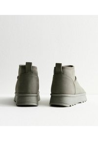 Botas de tobillo de ante gris con suela de goma gruesa, que presentan una lengüeta en el talón y sutiles detalles de costura, mostradas desde atrás.