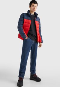 Chaqueta de plumas roja y azul marino con cremallera frontal, ambos colores en secciones horizontales, combinada con una camiseta negra y jeans azules. Calzado negro.