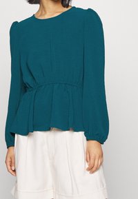 Blusa lunga teal a maniche lunghe con vita elastica, orlo peplum fluido e polsini arricciati, abbinata a pantaloni corti di colore chiaro.