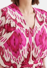 Klänning med rosa mönster som har en djup V-ringning, bred krage och mjuk tygtextur med ett abstrakt mönster i nyanser av rosa och kräm.