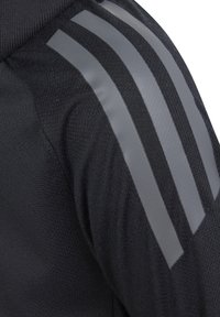 Veste de sport noire en tissu texturé avec trois rayures grises réfléchissantes le long de l'épaule. Design minimaliste pour la fonctionnalité.