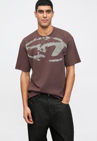 Mladý muž s krátkymi vlasmi, oblečený v hnedom voľnom tričku s abstraktným šedým potlačou a čiernych džínsoch, s jednou rukou v vrecku, na neutrálne pozadí.