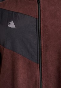 Half-zip fleecejack met bordeauxrode zachte stof en een zwarte textuurpanel met een wit driehoekig ontwerp op de borst.
