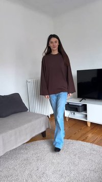 Femme en chemise marron à manches longues et jean bleu marchant sur un tapis rond gris dans un salon avec un canapé, un radiateur, une télévision et un sol en bois.