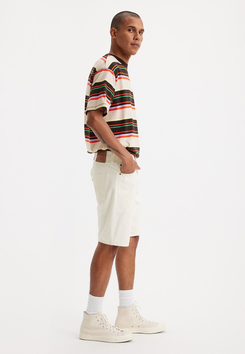 Kurzärmliges, gestreiftes T-Shirt in Beige, Grün, Rot und Braun. Kombiniert mit beigen Shorts und hellen High-Top Sneakers.