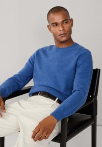 Blå crew-neck tröja i mjukt stickat tyg, kombinerad med cremefärgade byxor och ett brunt bälte, sittande på en svart stol mot en ljus bakgrund.