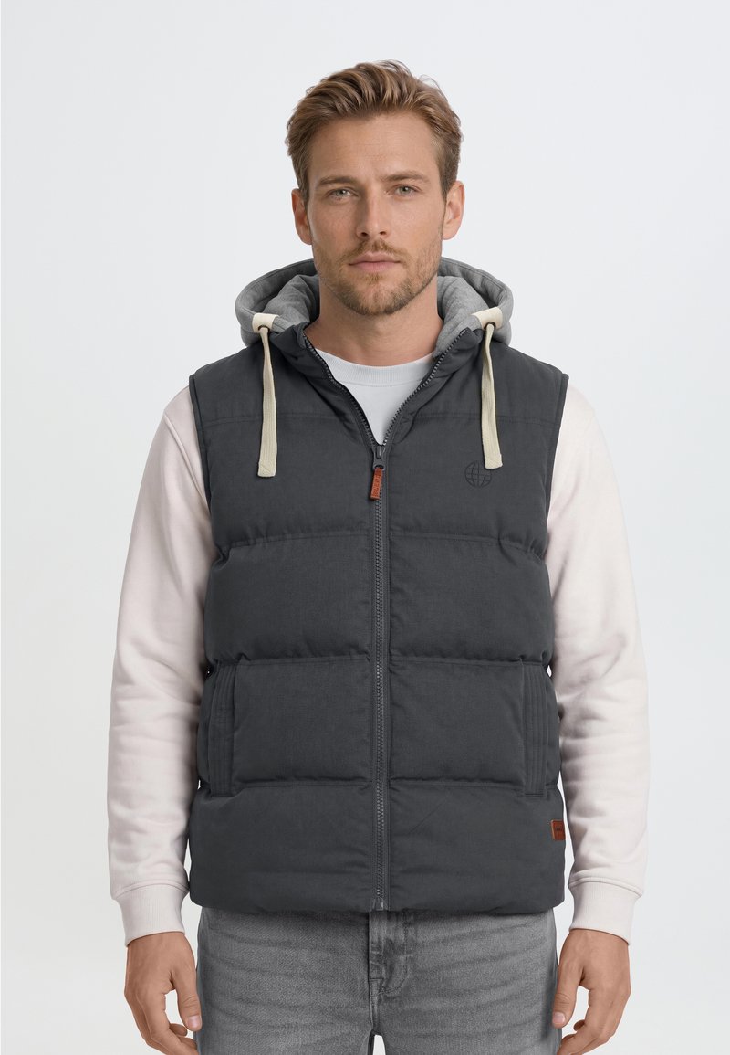 Hombre con chaleco acolchado negro con capucha sobre sudadera clara y vaqueros grises, de pie frente a un fondo claro y simple.