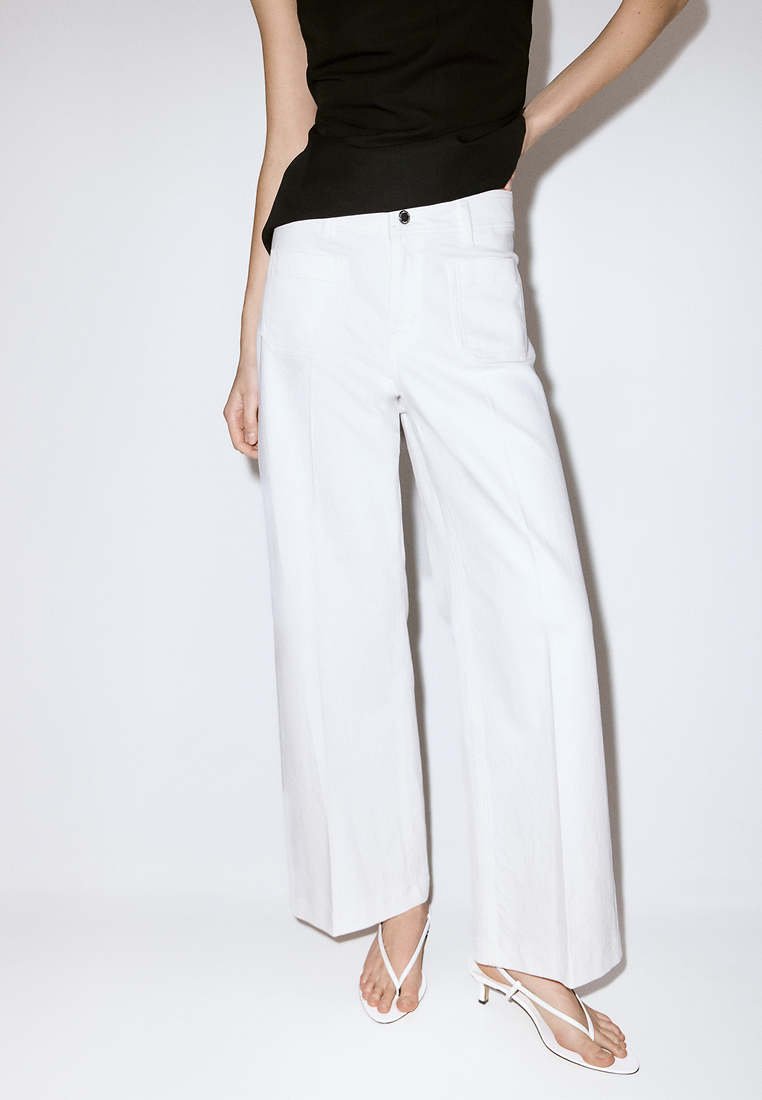 Femme portant un pantalon blanc à jambes larges, un top noir sans manches et des sandales blanches à talons avec des brides, debout devant un fond clair uni.