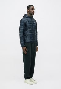 Tengerészkék steppelt dzseki vízszintes varrásokkal, cipzáras záródással és kapucnival. Sötétzöld melegítőnadrággal és törtfehér tornacipővel viselve.