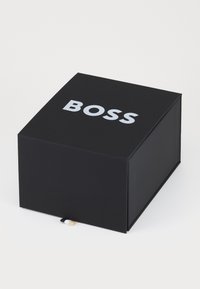 Boîte noire avec finition mate, présentant un grand logo blanc "BOSS" sur le dessus. Tirette latérale en noir avec un accent beige.