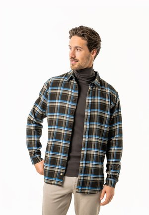 Mann trägt schwarzen Rollkragenpullover unter blau-braun kariertem Hemd mit beigen Hosen, schaut zur Seite, Hand in der Tasche, vor weißem Hintergrund.