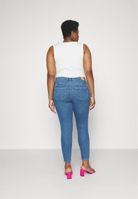 Klassiske blå denimjeans med baklommer og en lys tan-merke på livlinen, kombinert med en hvit ermeløs topp og rosa høyhælte sandaler.