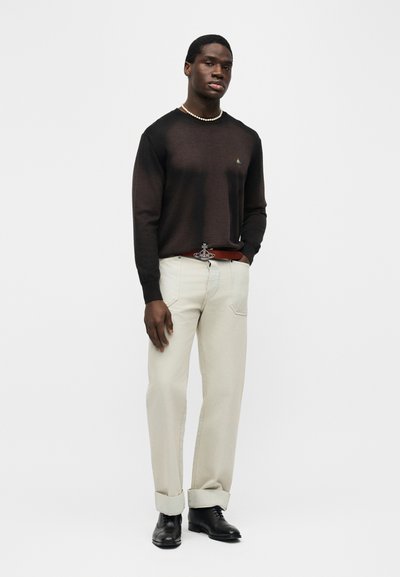 Άνδρας που φοράει διάφανο μαύρο πουκάμισο με μακριά μανίκια, περιδέραιο με πέρλες, μπεζ παντελόνι με μανσέτες και κόκκινη ζώνη, και μαύρα παπούτσια, στέκεται με το ένα χέρι στην τσέπη.