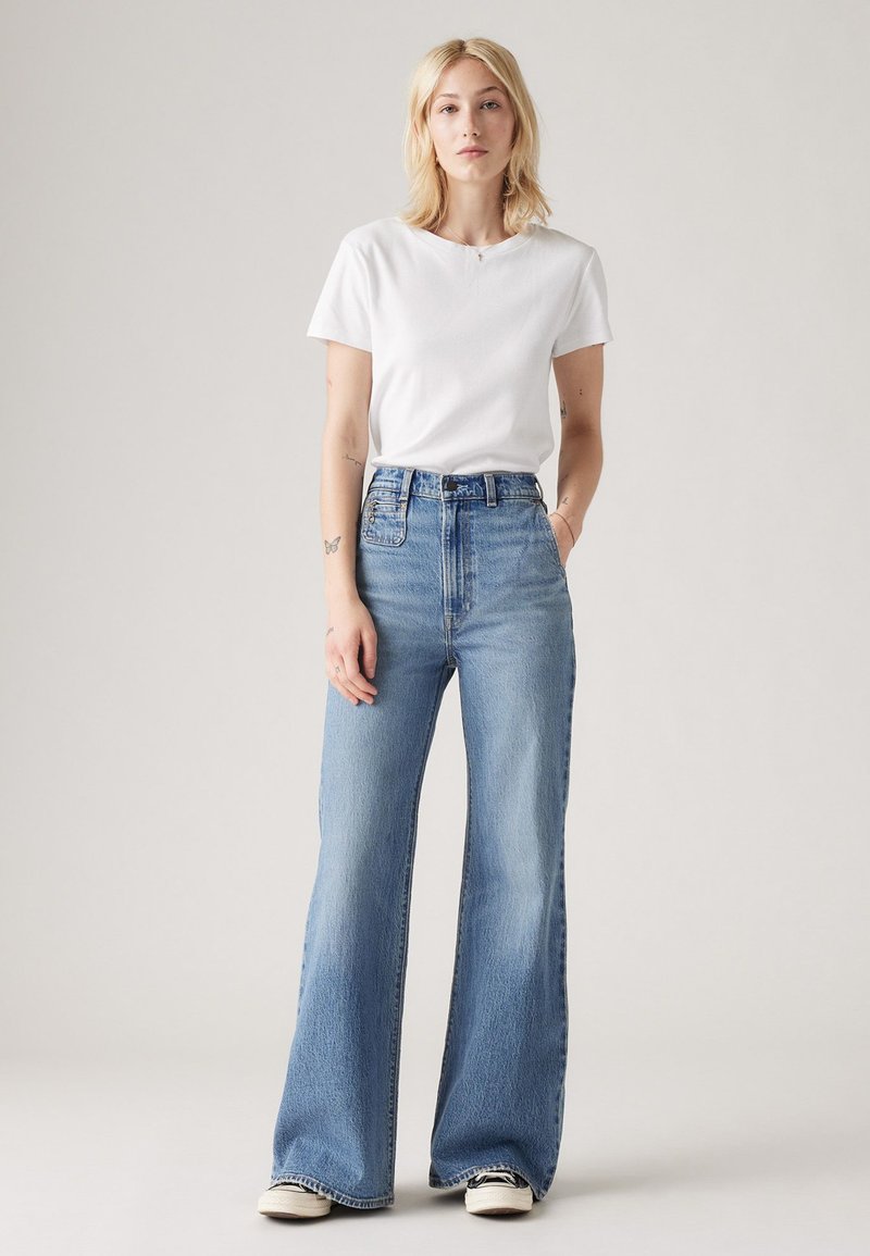 T-shirt en coton blanc associé à un jean flare en denim bleu clair taille haute, avec poches avant et décoloration subtile sur le tissu.