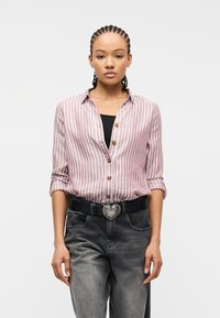 Νεαρή γυναίκα με πλεξούδες, που φορά ροζ και λευκό ριγέ πουκάμισο με κουμπιά, μαύρο τοπ, μαύρη ζώνη με καρδιόσχημη αγκράφα και ξεθωριασμένο τζιν.