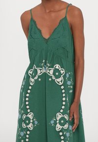 Zelené bezrukávové šaty s květinovou výšivkou v bílé a modré, nastavitelné ramínka a designem ve tvaru V. Krajkové detaily podél výstřihu.