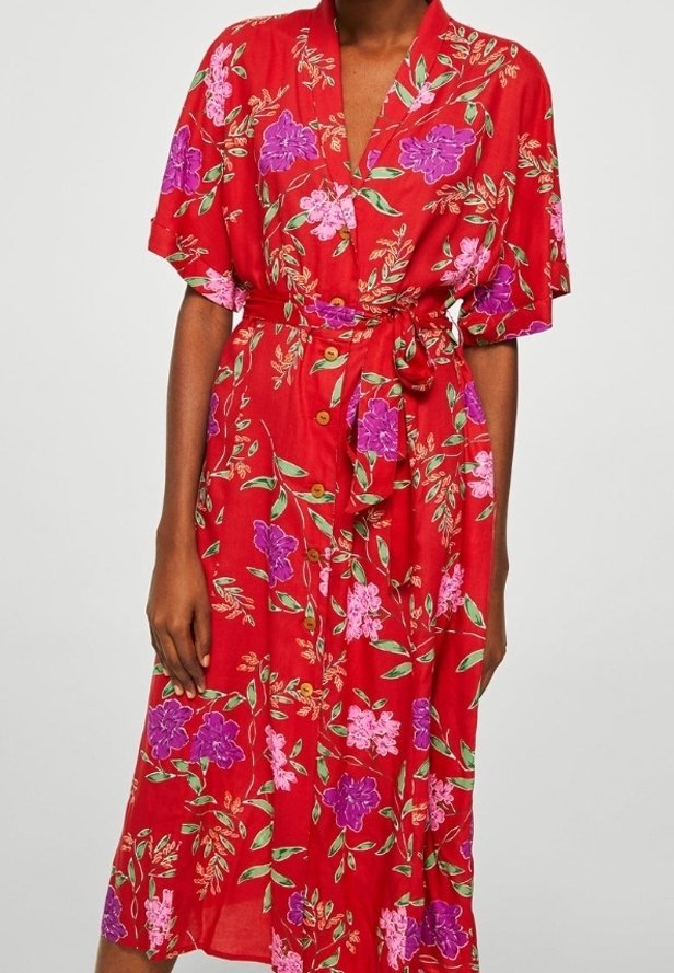 Femme portant une robe midi rouge à fleurs violettes et roses, manches courtes, boutonnée sur le devant, avec une ceinture nouée à la taille.