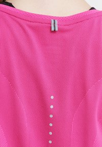 Rosa idrottstop med en texturerad tyg och ett subtilt nätmönster, en grå accentflik vid halsringningen och reflekterande prickar längs ryggen.