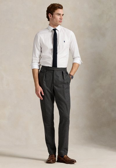 White dress shirt with button cuffs, dark tie, tailored gray trousers with pleats, and brown leather shoes. Model stands against a neutral background.