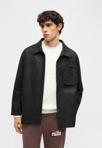 Νεαρός άνδρας που φοράει μαύρο σακάκι με κουμπιά και μεγάλη τσέπη στο στήθος με φερμουάρ, πάνω από λευκό πουκάμισο και καφέ φόρμα Nike.