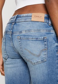 Jeans de denim azul claro con un diseño ajustado, que cuentan con una etiqueta de cuero con la palabra "ONLY" y costuras naranjas en los bolsillos. Vista trasera.