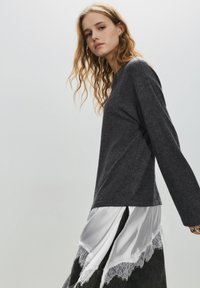 Mørkegrå langærmet top med løs pasform, lagdelt med hvid satin og blonder ved kanten, med sideslids for øget bevægelse.
