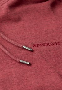 Tela de sudadera roja con textura, con extremos de cordón en tono plateado y logotipo bordado "SUPERDRY" en un rojo ligeramente más oscuro.