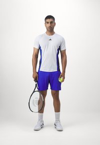 Witte sportshirt met blauwe en zwarte accenten, paarse shorts, houdt een tennisracket en bal vast, draagt witte schoenen met zwarte strepen.