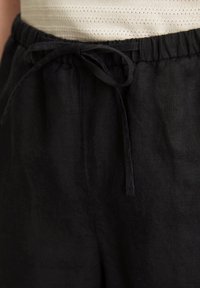 Schwarze Leinenhose mit einem Bund zum Zuziehen, weichem Material und lockerer Passform, teilweise sichtbar über einem hellen Oberteil.